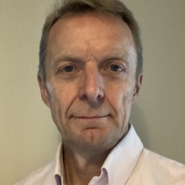 A middle-aged man with short light brown hair, wearing a light-colored collared shirt, faces the camera against a plain background.
