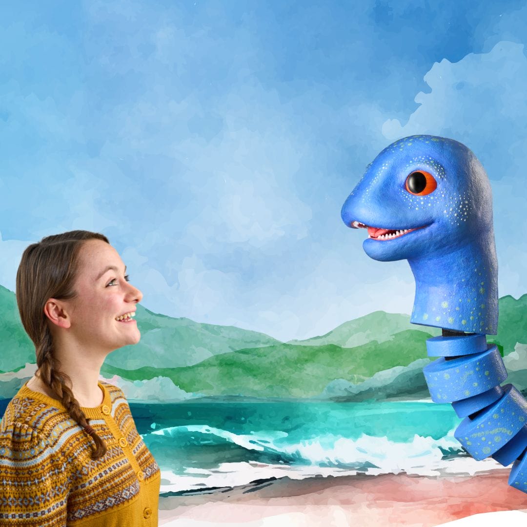 A woman in a yellow sweater smiles at a large blue puppet resembling a sea creature, with an illustrated ocean and mountains in the background.