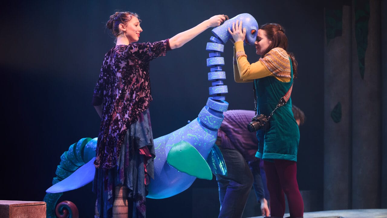 Two women interact with a large blue sea creature puppet on stage during a theatrical performance.