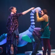 Two women interact with a large blue sea creature puppet on stage during a theatrical performance.