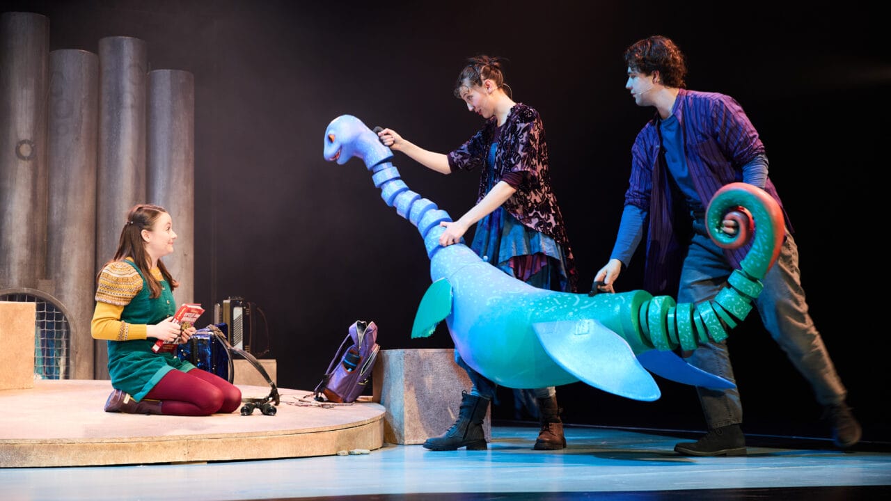 Three actors on stage interact with a large, blue puppet during a theatrical performance with minimal set pieces in the background.