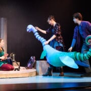 Three actors on stage interact with a large, blue puppet during a theatrical performance with minimal set pieces in the background.