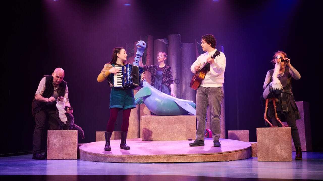 Five performers on stage with puppets; two play musical instruments, one stands behind a large blue puppet, and others operate smaller animal puppets. Stage is lit with purple and pink lighting.