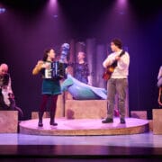 Five performers on stage with puppets; two play musical instruments, one stands behind a large blue puppet, and others operate smaller animal puppets. Stage is lit with purple and pink lighting.