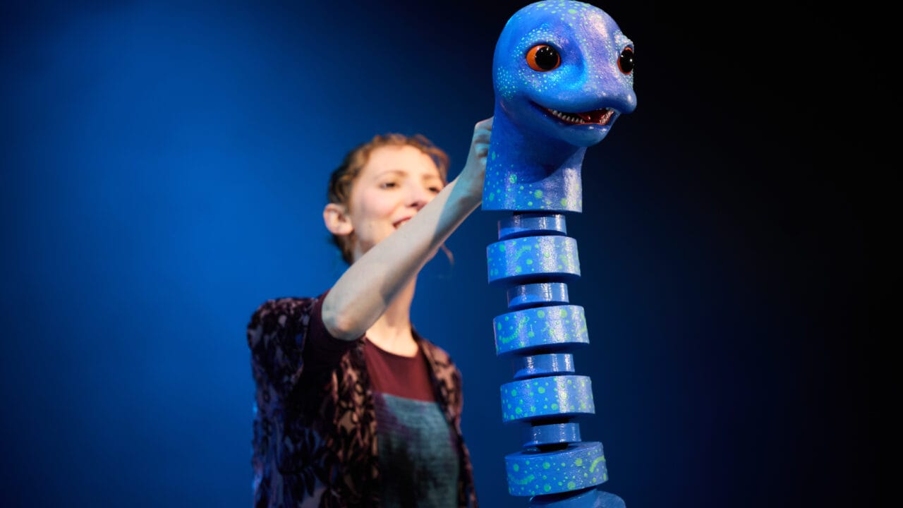 A person operates a segmented blue puppet resembling a loch ness monster against a blue background.