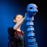 A person operates a segmented blue puppet resembling a loch ness monster against a blue background.