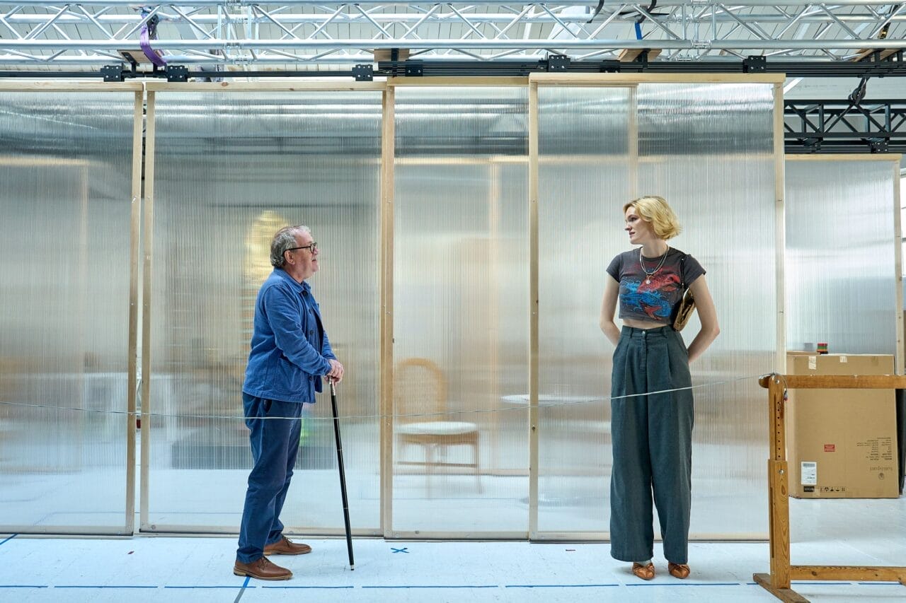 An older man with a cane stands facing a tall woman with short blond hair, in front of a transparent partition in a modern indoor setting, reminiscent of an elegant scene from Death on the Nile.