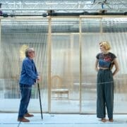 An older man with a cane stands facing a tall woman with short blond hair, in front of a transparent partition in a modern indoor setting, reminiscent of an elegant scene from Death on the Nile.