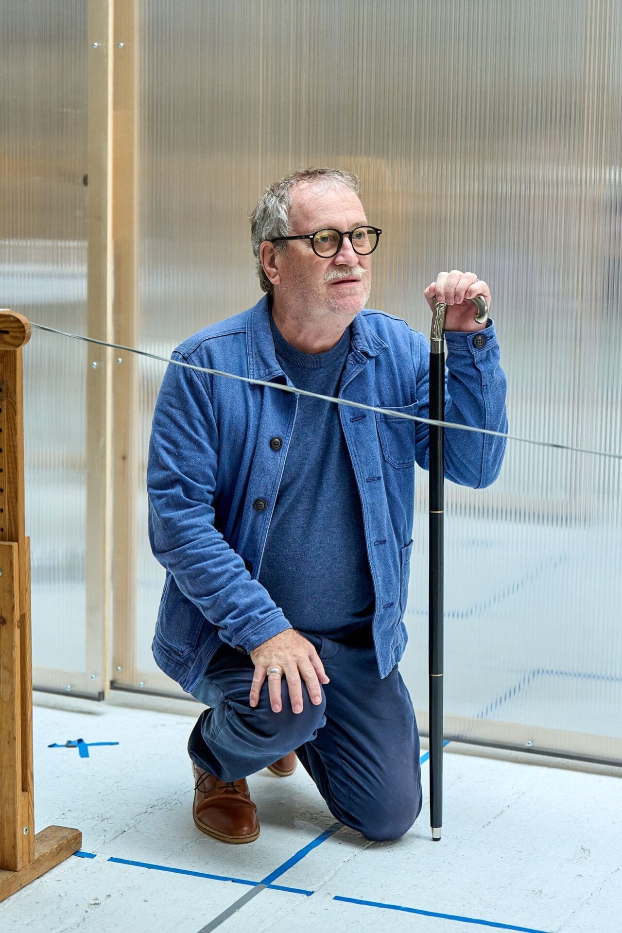 A man in glasses and a blue outfit kneels on one knee indoors, holding a black cane upright beside a taut wire and wooden object, evoking the suspenseful atmosphere of Death on the Nile.