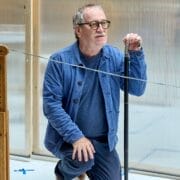 A man in glasses and a blue outfit kneels on one knee indoors, holding a black cane upright beside a taut wire and wooden object, evoking the suspenseful atmosphere of Death on the Nile.