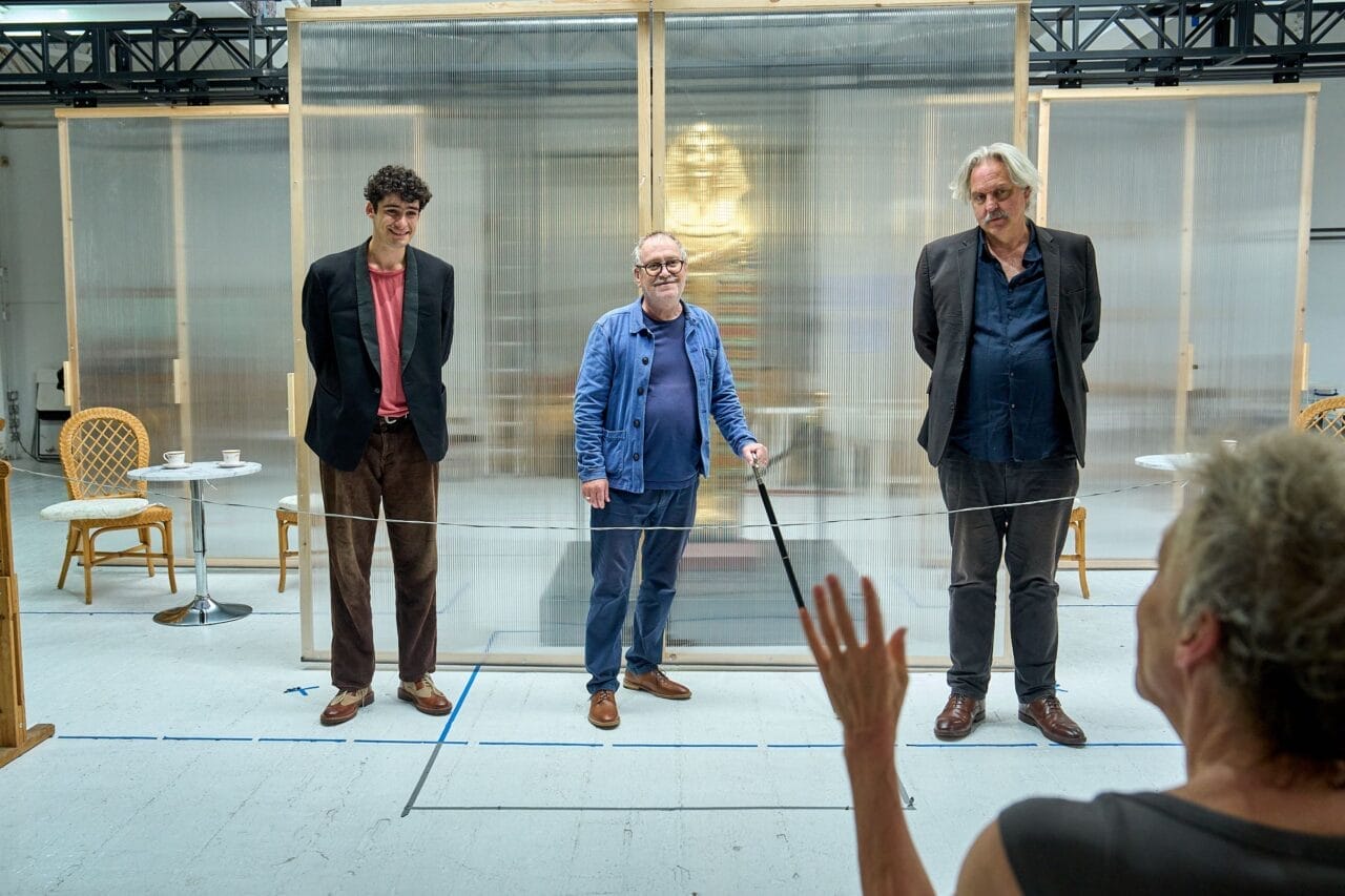 Three men stand in a row facing a seated person who gestures towards them, inside a room with chairs, tables, and a translucent backdrop—evoking the suspenseful atmosphere of Death on the Nile.