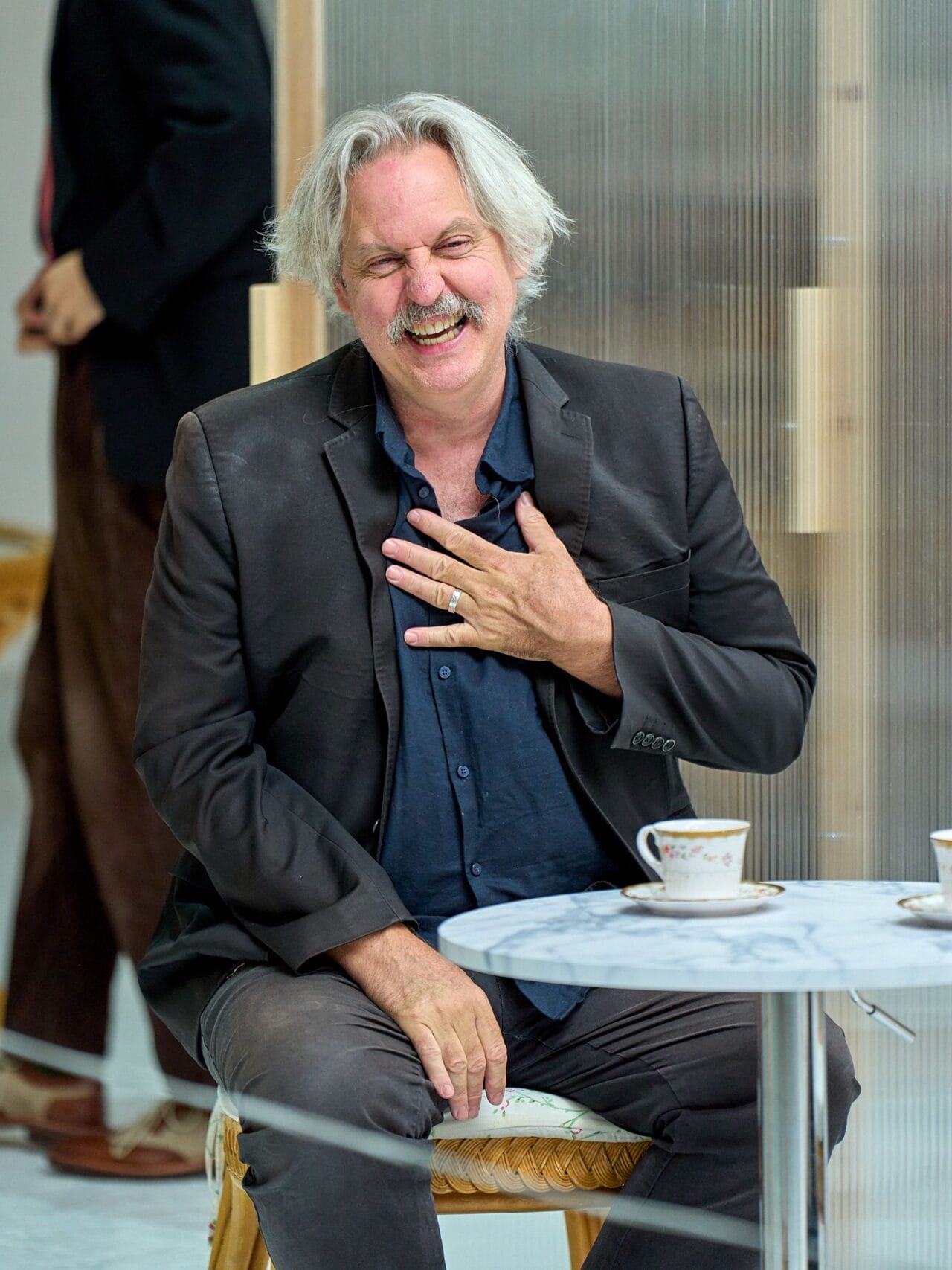 A middle-aged man with gray hair and a mustache sits at a table, smiling and touching his chest, evoking the charm of Death on the Nile. Teacups and saucers are neatly arranged in front of him.