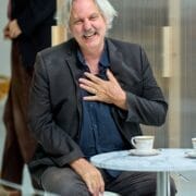 A middle-aged man with gray hair and a mustache sits at a table, smiling and touching his chest, evoking the charm of Death on the Nile. Teacups and saucers are neatly arranged in front of him.