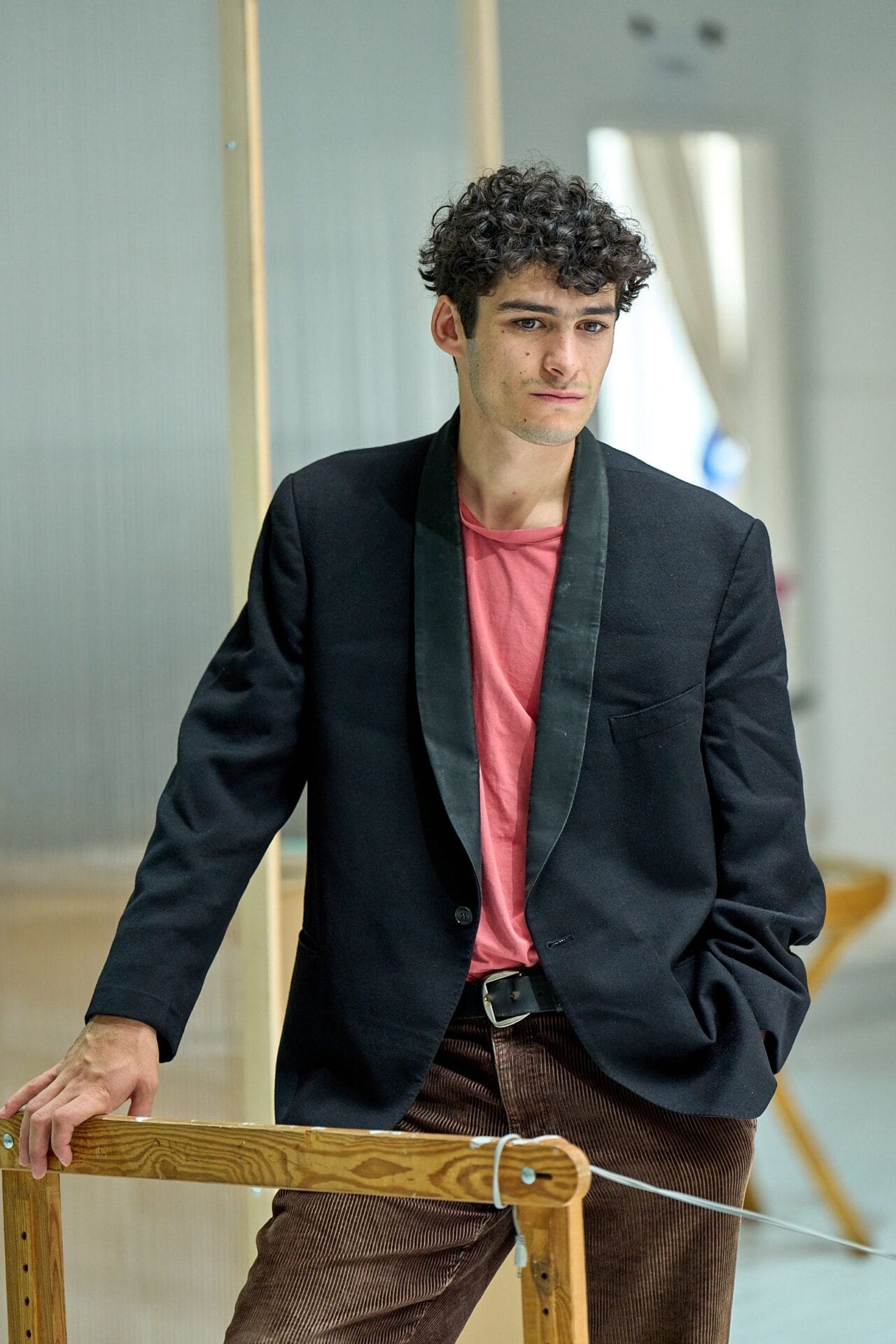A man with curly hair wearing a black blazer, pink shirt, and brown corduroy pants stands indoors, evoking a Death on the Nile vibe as he leans on a wooden structure with one hand in his pocket.
