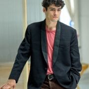 A man with curly hair wearing a black blazer, pink shirt, and brown corduroy pants stands indoors, evoking a Death on the Nile vibe as he leans on a wooden structure with one hand in his pocket.