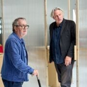 Two men stand indoors near a translucent partition; one holds a cane. Both appear to be engaged in conversation or observing something, evoking a scene reminiscent of Death on the Nile.
