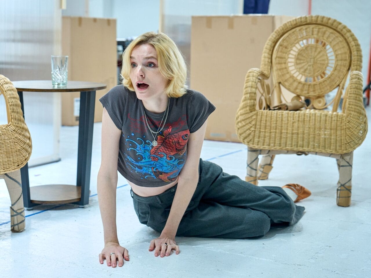 A person with short blond hair, wearing a printed t-shirt and loose pants, kneels on the floor indoors between two wicker chairs and a small table, evoking the relaxed yet mysterious atmosphere of Death on the Nile.