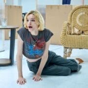 A person with short blond hair, wearing a printed t-shirt and loose pants, kneels on the floor indoors between two wicker chairs and a small table, evoking the relaxed yet mysterious atmosphere of Death on the Nile.