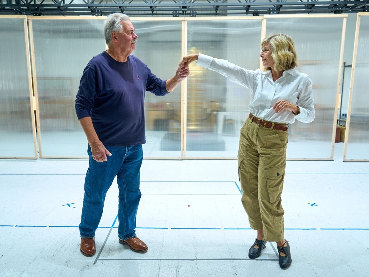 An older man and woman stand facing each other indoors; the woman lifts her arm while the man lightly holds her hand, evoking a graceful moment reminiscent of a scene from Death on the Nile as they practice a dance move.