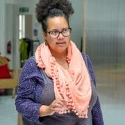 A woman with curly hair, glasses, and a pink scarf stands indoors in a blue polka-dot shirt and grey overalls, her thoughtful gaze to the side reminiscent of a character from Death on the Nile.