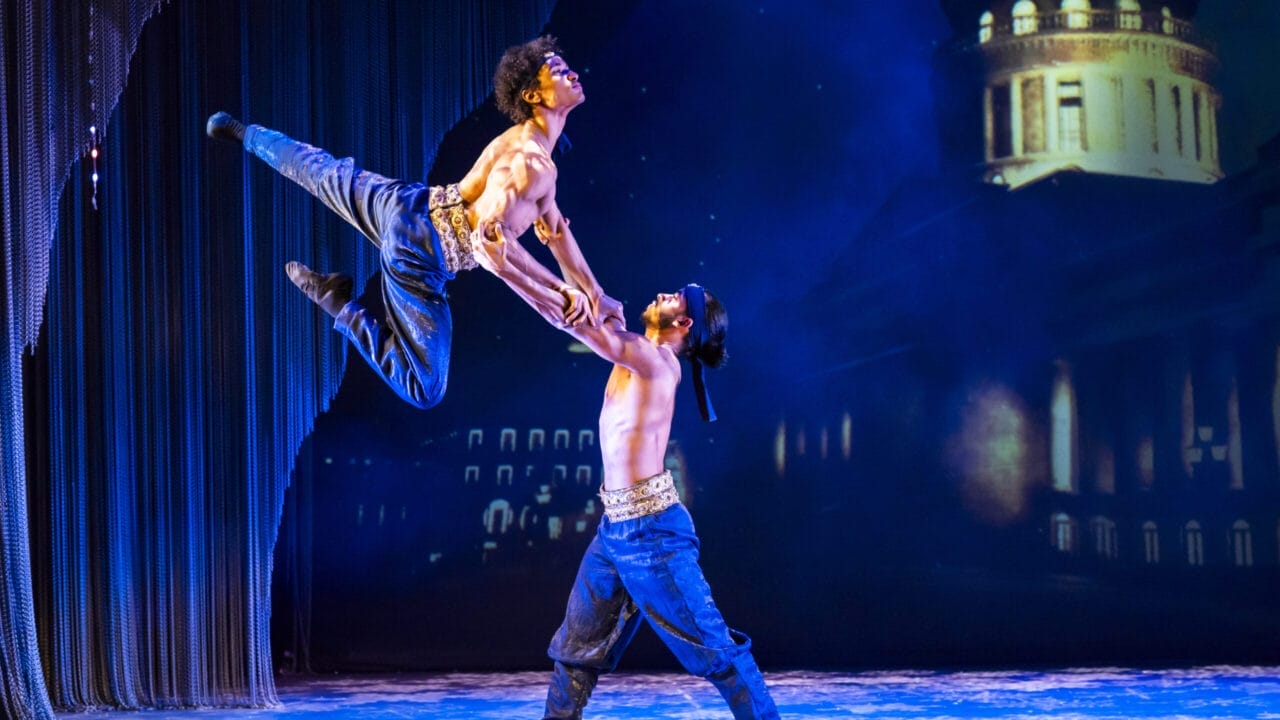 Two male dancers perform an acrobatic lift on stage, wearing jeans and belts, with a nighttime Havana cityscape projected in the background.