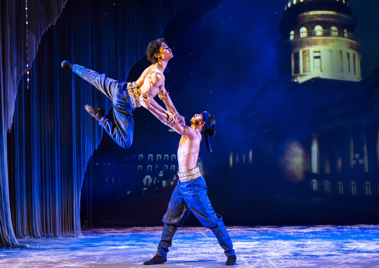 Two male dancers perform an acrobatic lift on stage, wearing jeans and belts, with a nighttime Havana cityscape projected in the background.