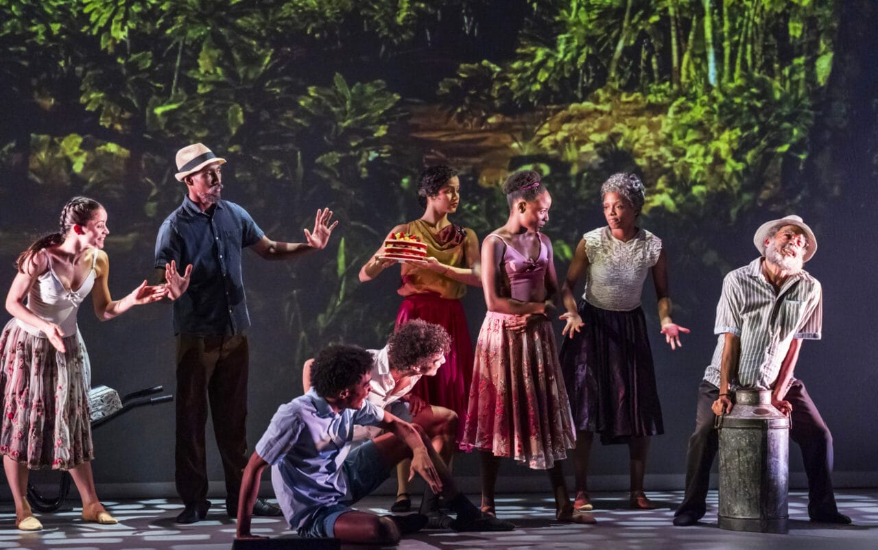 A group of people in colorful clothing perform on stage with a vibrant jungle backdrop; some stand, some sit, one holds a cake, and another leans on a metal drum, evoking the lively spirit of Havana.