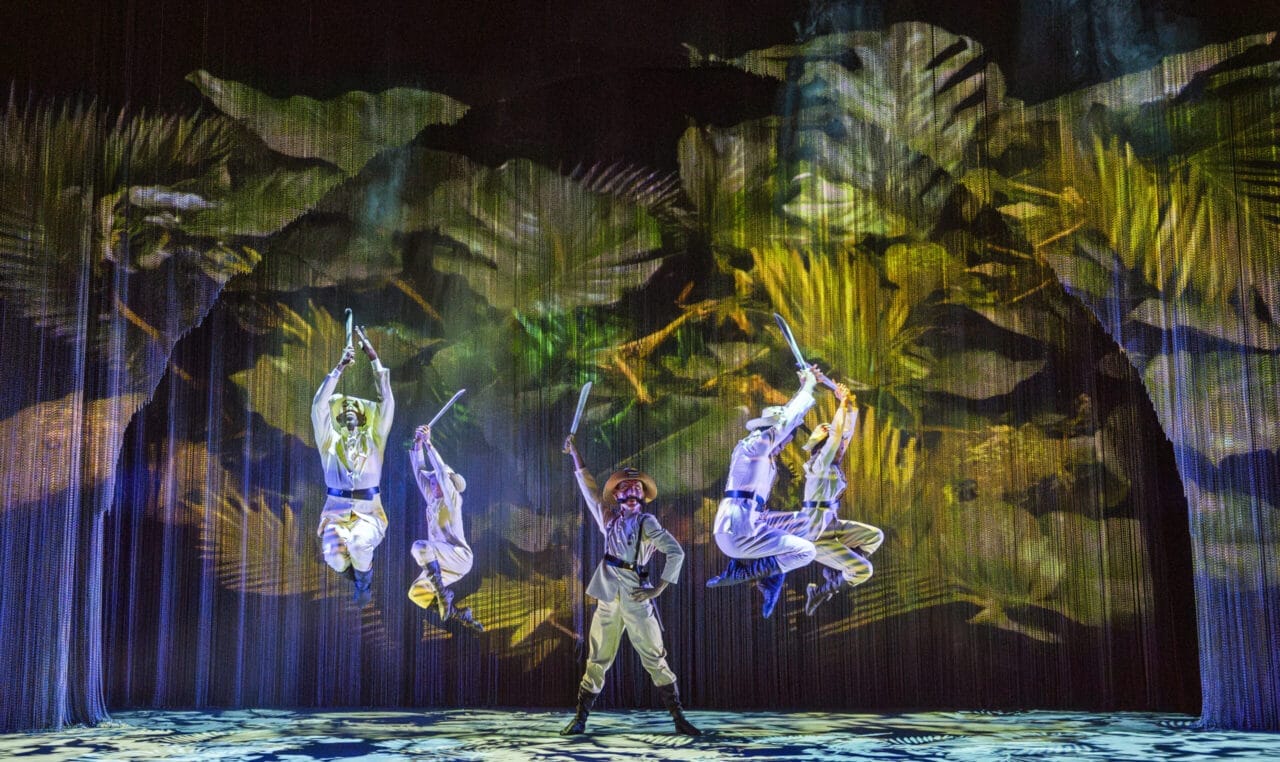 Five performers in jungle-themed costumes leap and pose with swords on stage, set against a vibrant rainforest backdrop—a thrilling twist inspired by Havana energy and the adventurous spirit of The Nutcracker.