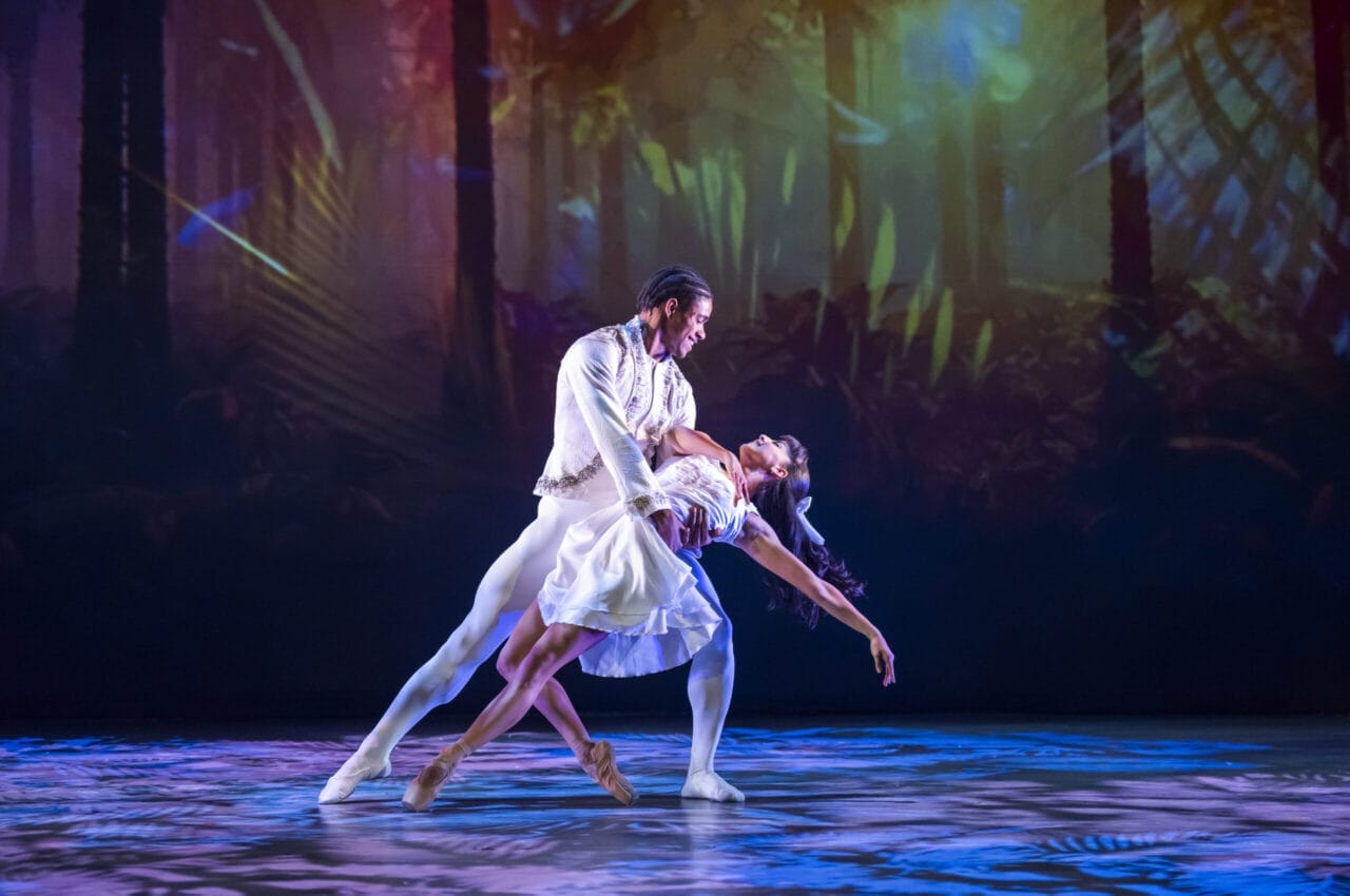 Two ballet dancers perform a dramatic pose on stage, with the male dancer supporting the female dancer in a dip. A colorful Nutcracker-inspired forest-themed backdrop is projected behind them.