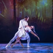 Two ballet dancers perform a dramatic pose on stage, with the male dancer supporting the female dancer in a dip. A colorful Nutcracker-inspired forest-themed backdrop is projected behind them.