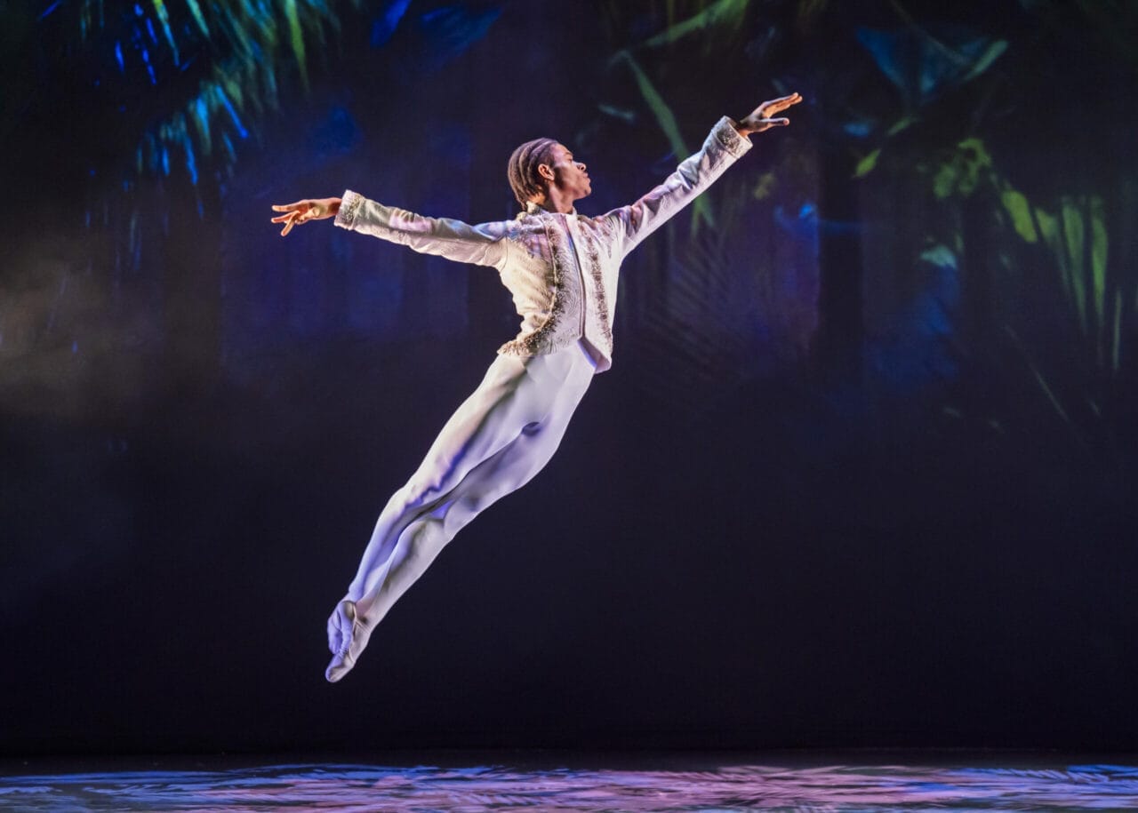 A male ballet dancer in a white costume performs a high leap on stage, capturing the spirit of Havana against a dark, colorful background reminiscent of The Nutcracker.