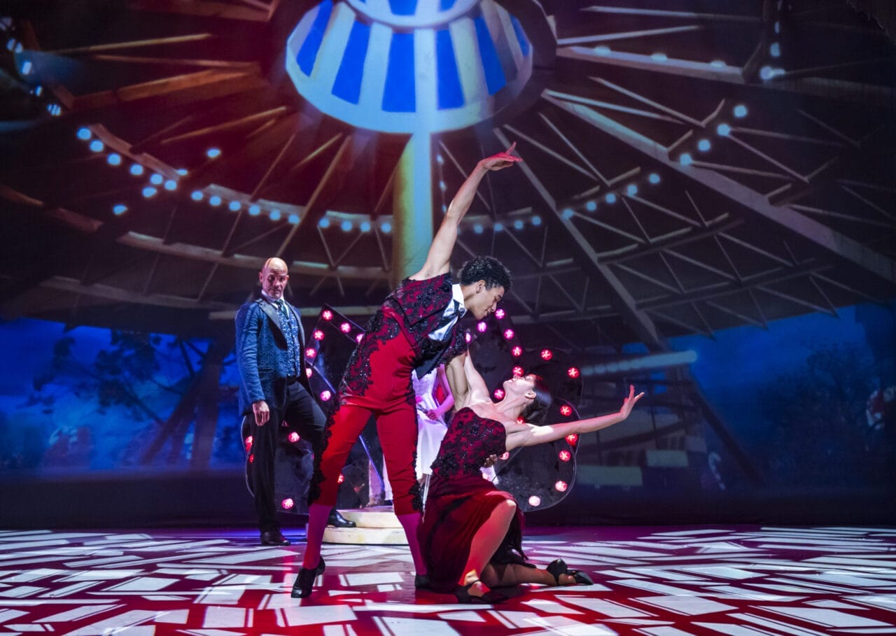 Two dancers perform an expressive pose on stage under dramatic lighting during a Nutcracker scene, while a third person stands in the background watching.