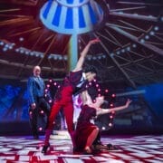 Two dancers perform an expressive pose on stage under dramatic lighting during a Nutcracker scene, while a third person stands in the background watching.