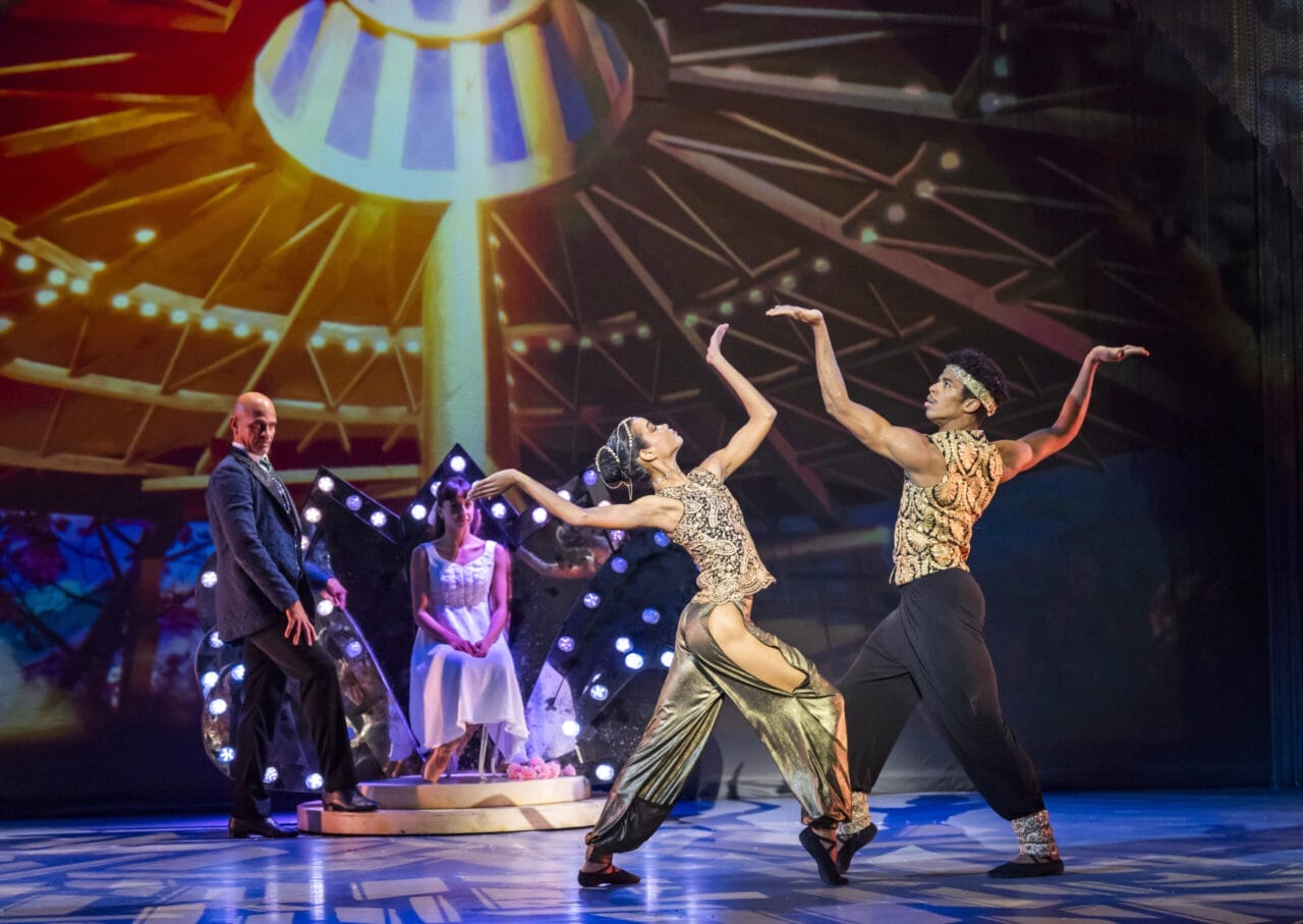 Four performers on stage; two in elaborate Nutcracker dance costumes pose mid-dance, while one sits on a bench and another stands, set against a colorful, illuminated Havana-inspired backdrop.