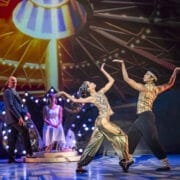 Four performers on stage; two in elaborate Nutcracker dance costumes pose mid-dance, while one sits on a bench and another stands, set against a colorful, illuminated Havana-inspired backdrop.