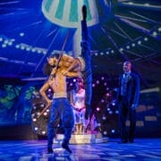 Three performers in elaborate Nutcracker-inspired costumes are on stage; one is balanced on another's shoulder with a dramatic leg extension, while a fourth person in a suit watches from the background, adding a touch of Havana elegance.