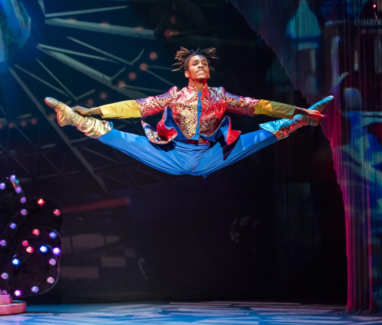 A performer in a colorful costume executes a high, mid-air split leap on stage under bright, theatrical lighting, reminiscent of the vibrant energy of Havana or a scene from the Nutcracker.