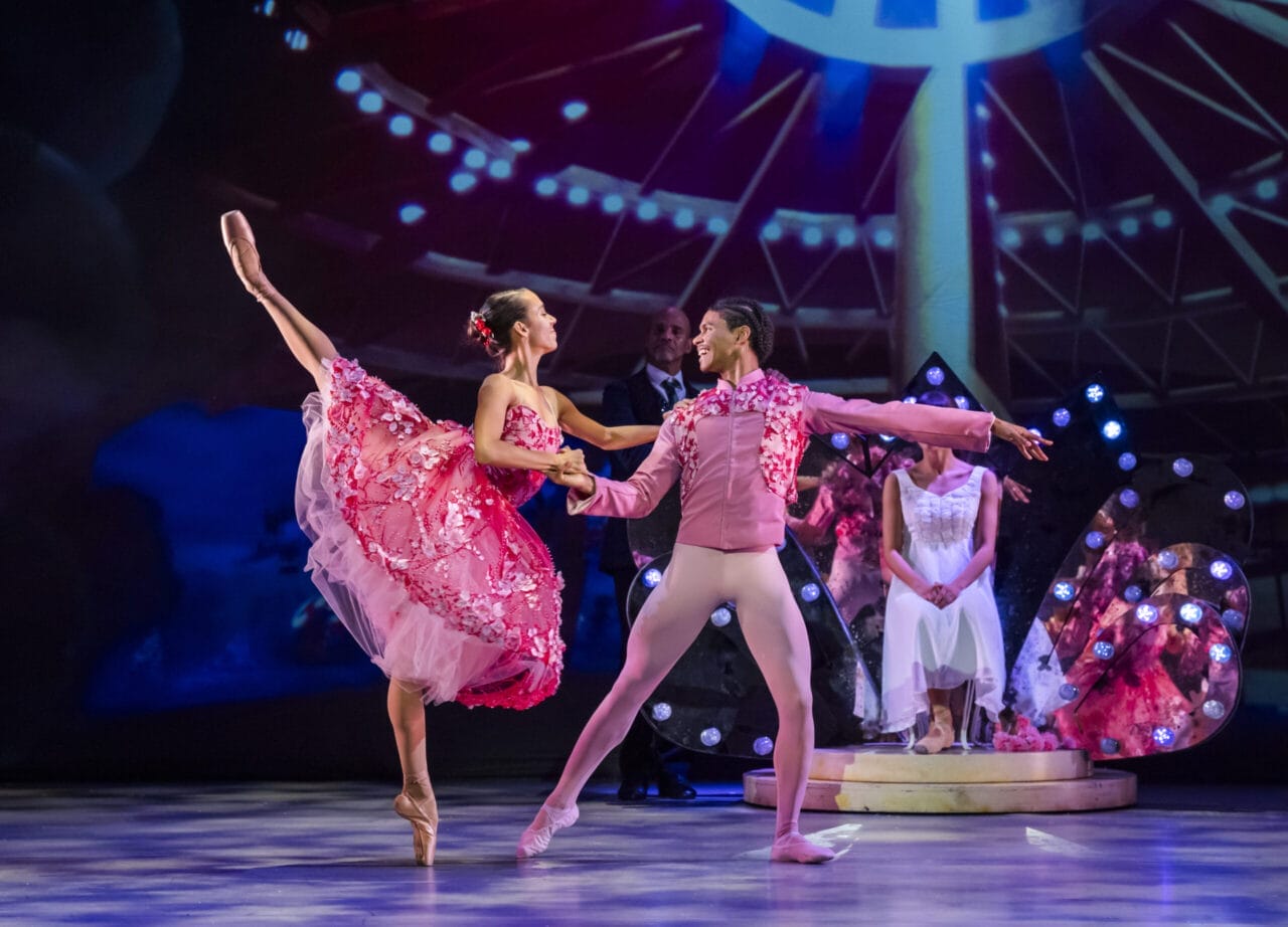 Two ballet dancers perform on stage in pink costumes, evoking scenes from the Nutcracker, with one dancer balancing on one leg and the other supporting. Stage lights and background props are visible.