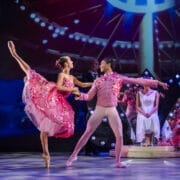 Two ballet dancers perform on stage in pink costumes, evoking scenes from the Nutcracker, with one dancer balancing on one leg and the other supporting. Stage lights and background props are visible.