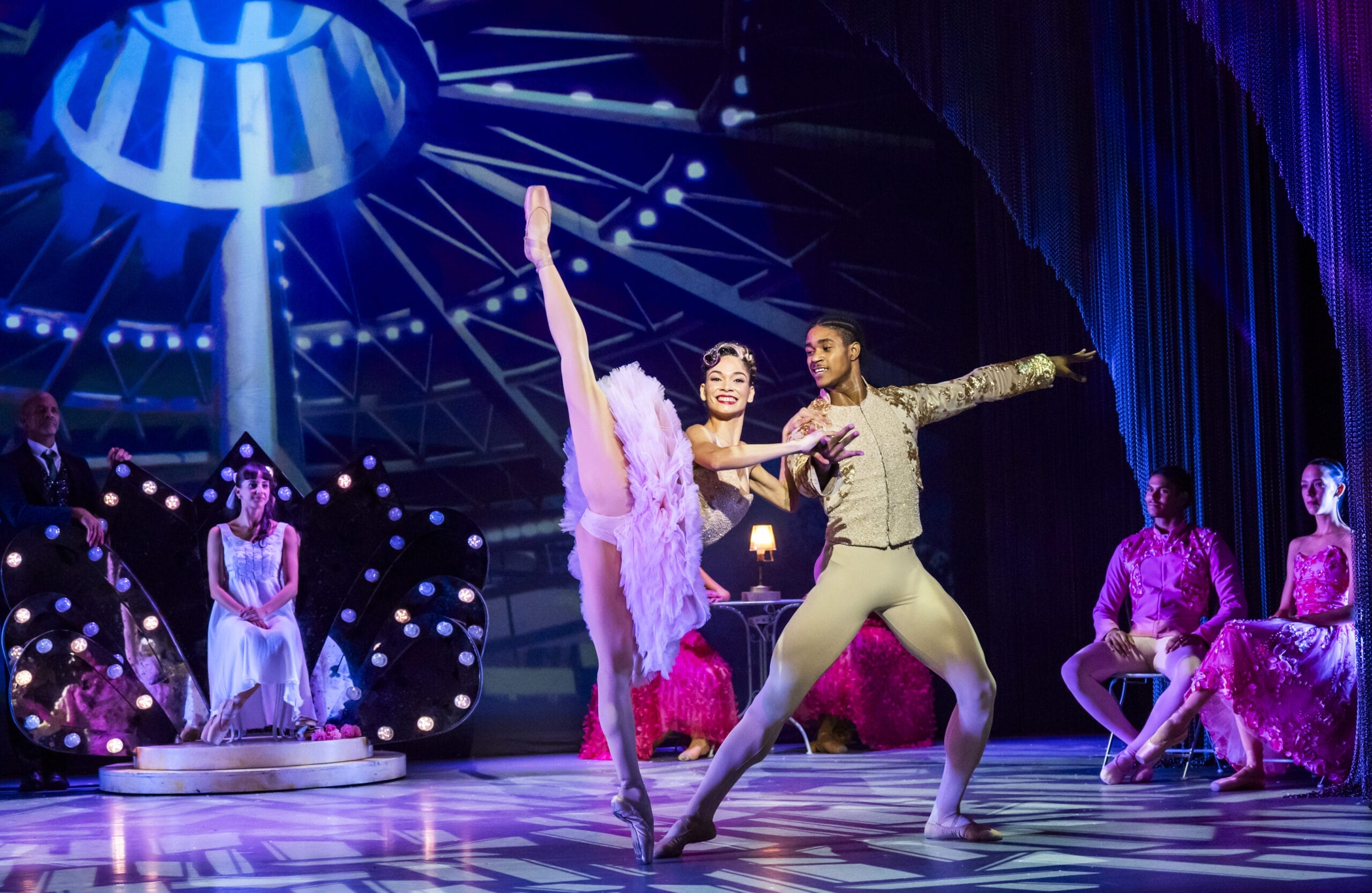 Two ballet dancers perform on stage in colorful Nutcracker costumes, with other performers seated in the background under dramatic lighting and festive stage decor.