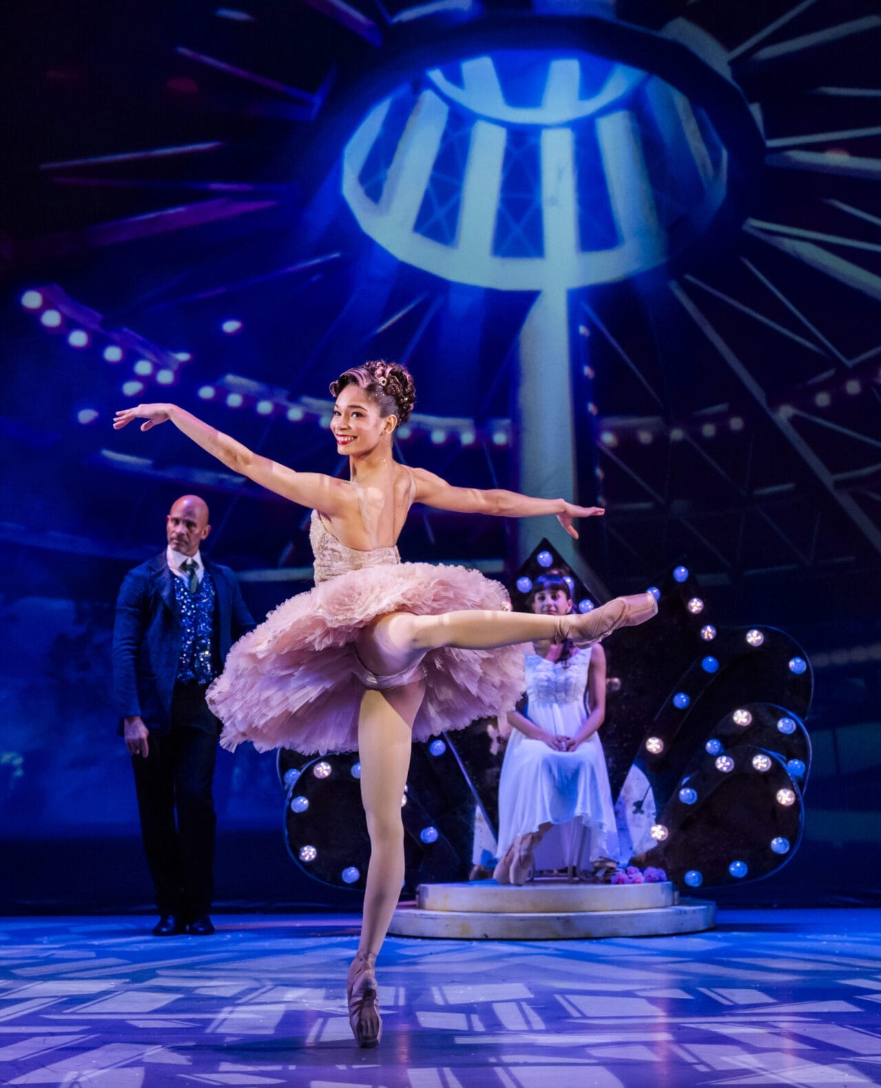 A ballerina in a pink tutu performs a Nutcracker ballet pose on stage, with a man standing and a seated woman in the background under bright stage lights.