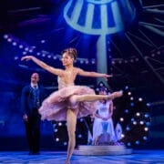 A ballerina in a pink tutu performs a Nutcracker ballet pose on stage, with a man standing and a seated woman in the background under bright stage lights.