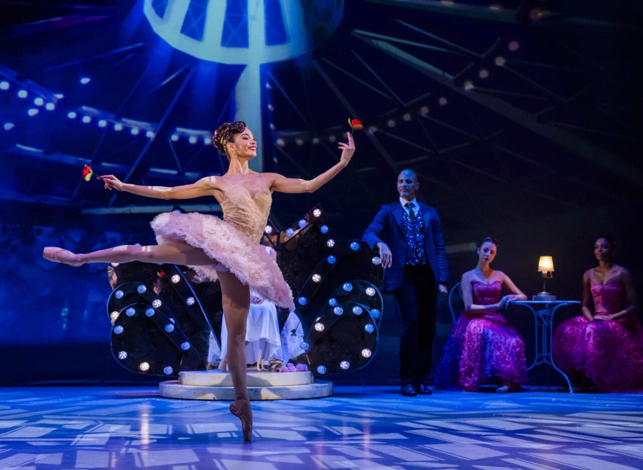 A ballerina in a pink tutu dances on stage holding red flowers, evoking the grace of the Nutcracker, with three people seated in the background under dramatic stage lighting.