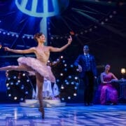 A ballerina in a pink tutu dances on stage holding red flowers, evoking the grace of the Nutcracker, with three people seated in the background under dramatic stage lighting.