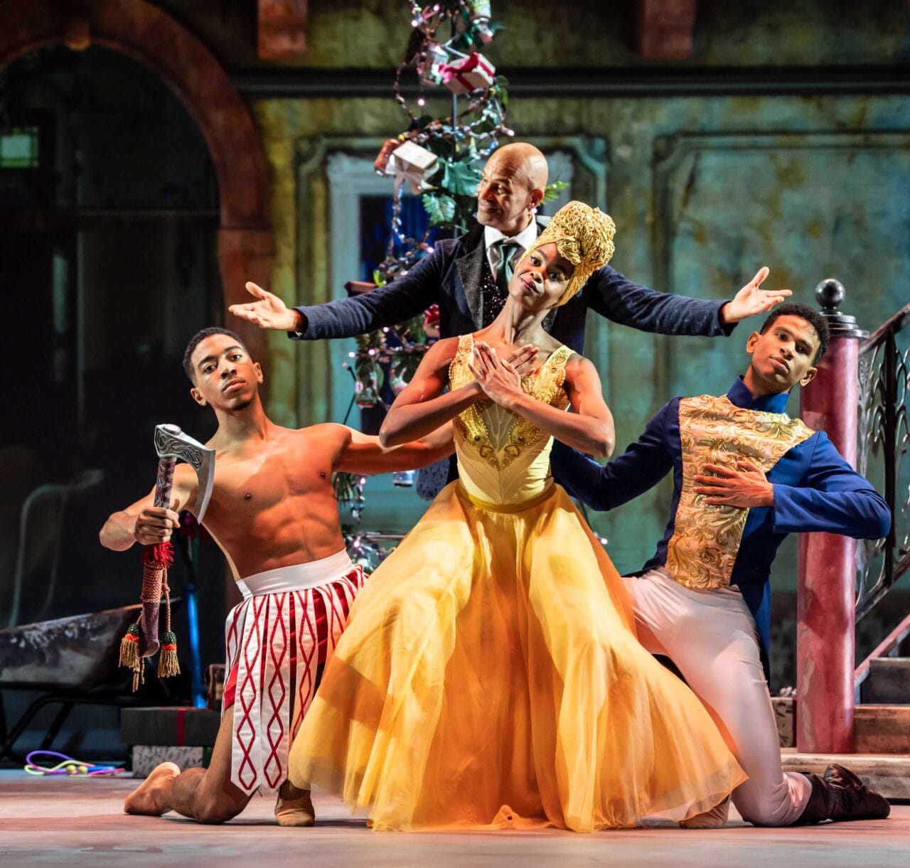 Three ballet dancers pose on stage in colorful costumes; one wears a yellow dress, one is shirtless with red-patterned pants, and another wears blue and gold, with a suited figure standing behind them.