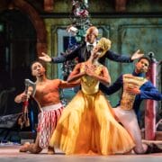 Three ballet dancers pose on stage in colorful costumes; one wears a yellow dress, one is shirtless with red-patterned pants, and another wears blue and gold, with a suited figure standing behind them.