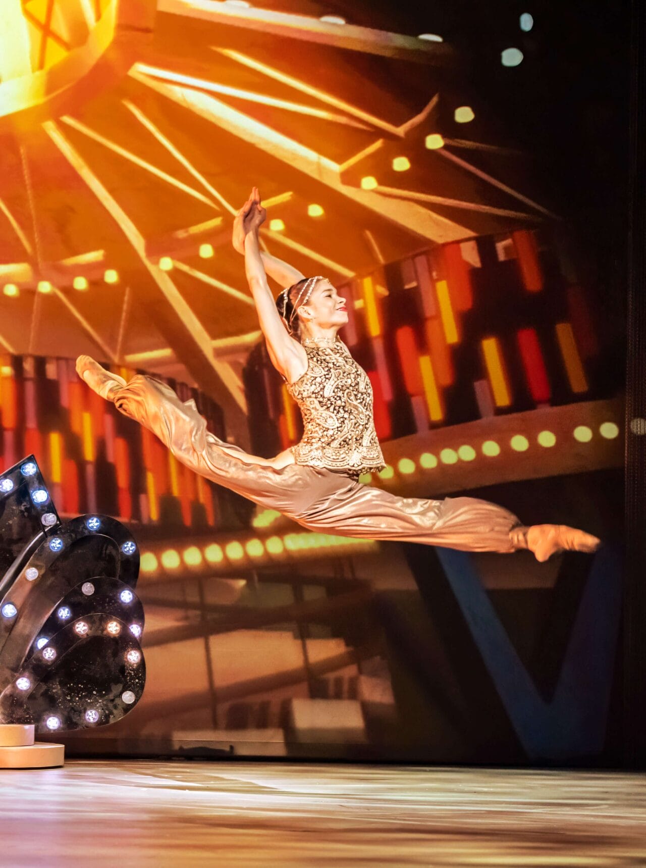 A ballet dancer in a gold costume performs a grand jeté on stage, with bright theater lights and a colorful background behind her.