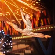A ballet dancer in a gold costume performs a grand jeté on stage, with bright theater lights and a colorful background behind her.