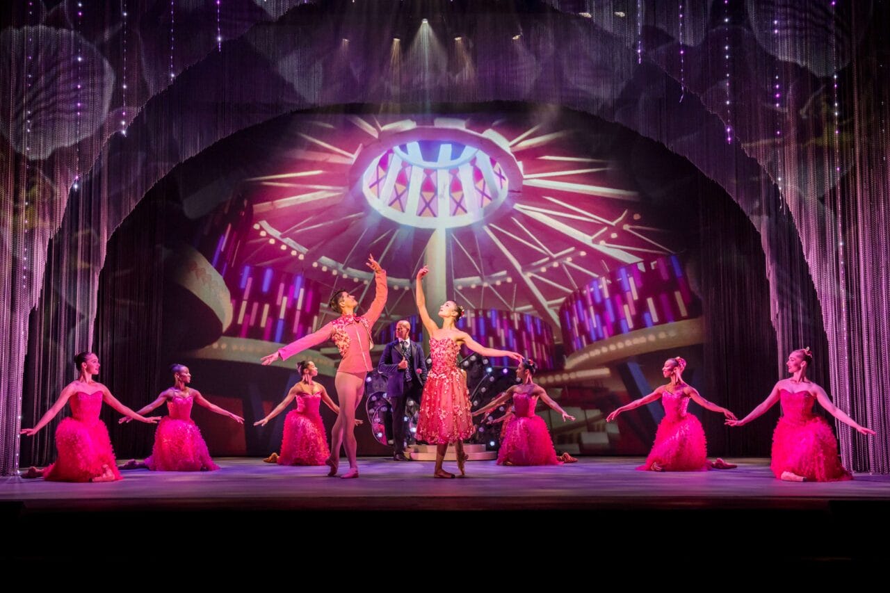 Ballet dancers in red costumes perform on a stage with dramatic lighting and a circular art deco backdrop, surrounded by glittering curtains.