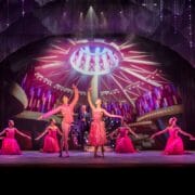 Ballet dancers in red costumes perform on a stage with dramatic lighting and a circular art deco backdrop, surrounded by glittering curtains.